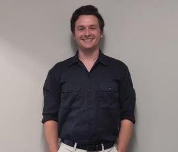 Male employee with brown hair wearing a black shirt and khakis smiling in front of a plain background. 
